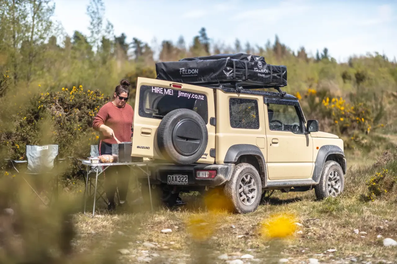 White Suzuki Jimny with rooftop tent in golden hour, mountain backdrop. – Jimny Rentals.
