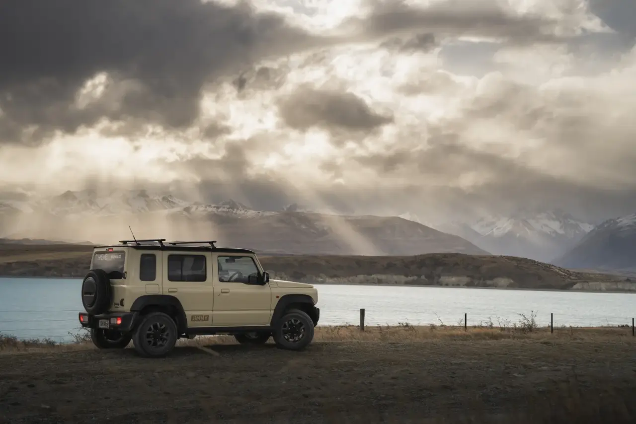 Suzuki Jimny driving on winding road with snow-capped mountains. – Jimny Rentals.