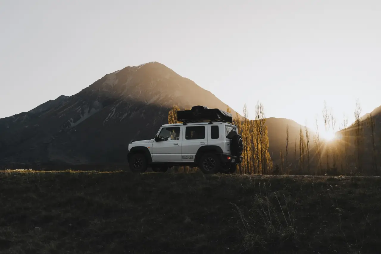 Suzuki Jimny parked by water with mountain backdrop. – Jimny Rentals.