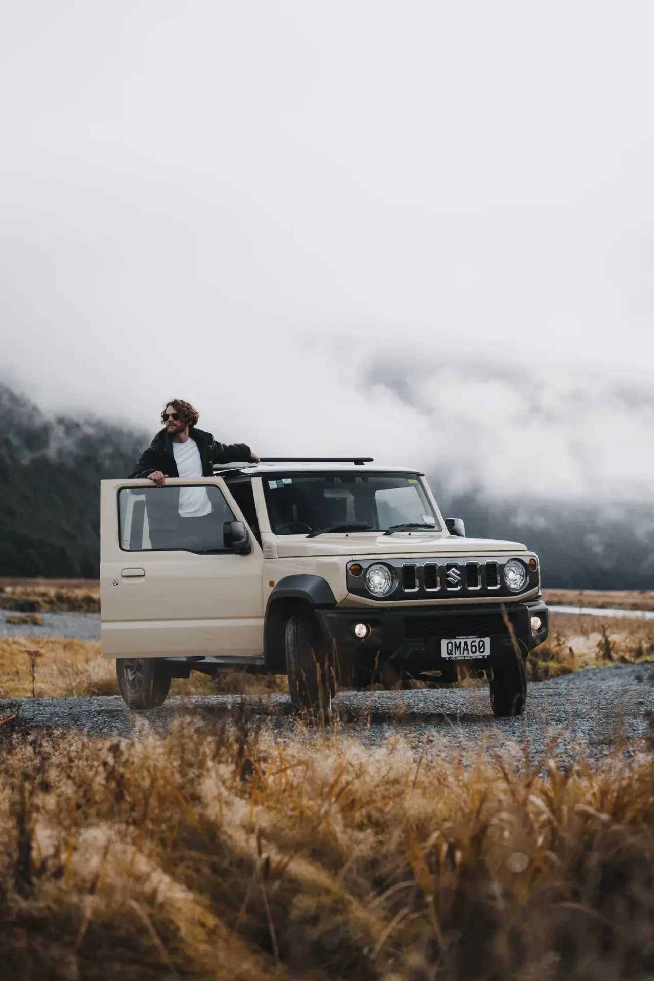 Suzuki Jimny beside lake with dramatic mountain and cloudscape. – Jimny Rentals.