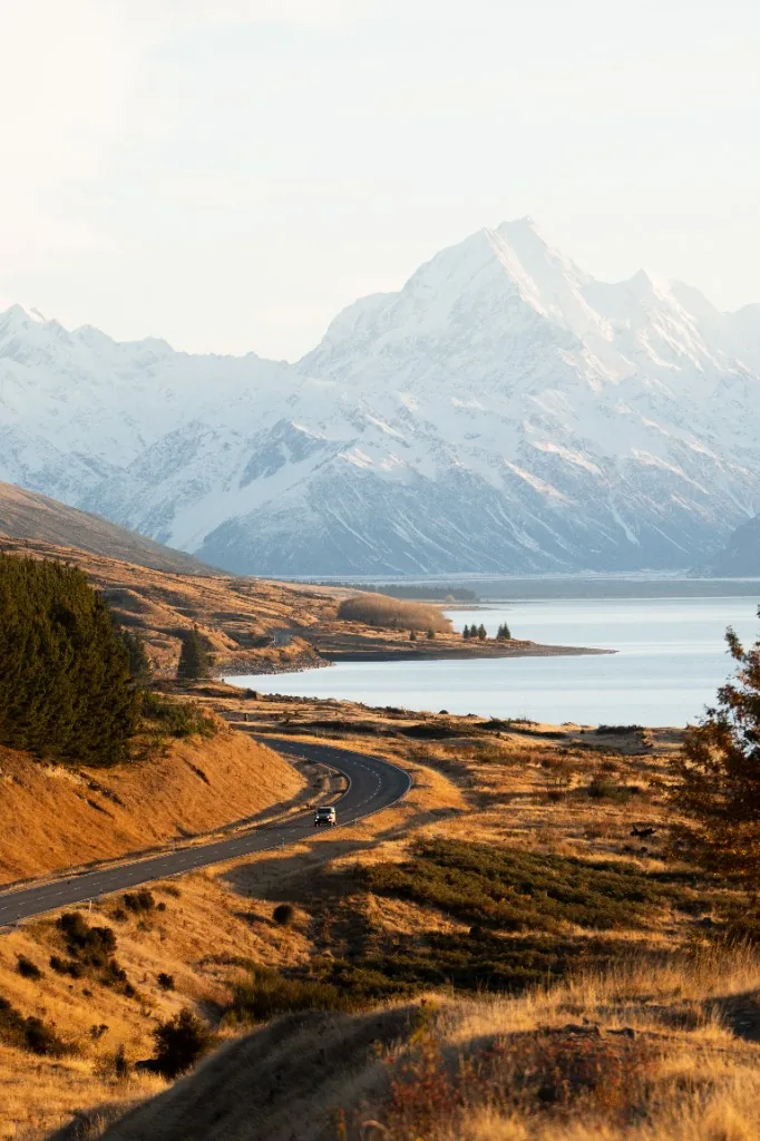 White Suzuki Jimny on a snowy South Island road at dusk with headlights and falling snow.