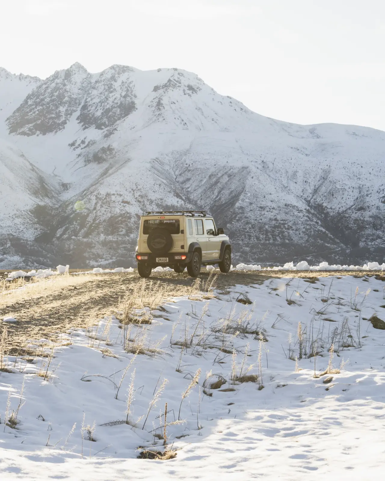 Beige Suzuki Jimny with headlights on in alpine setting, snow-capped mountains. – Jimny Rentals.