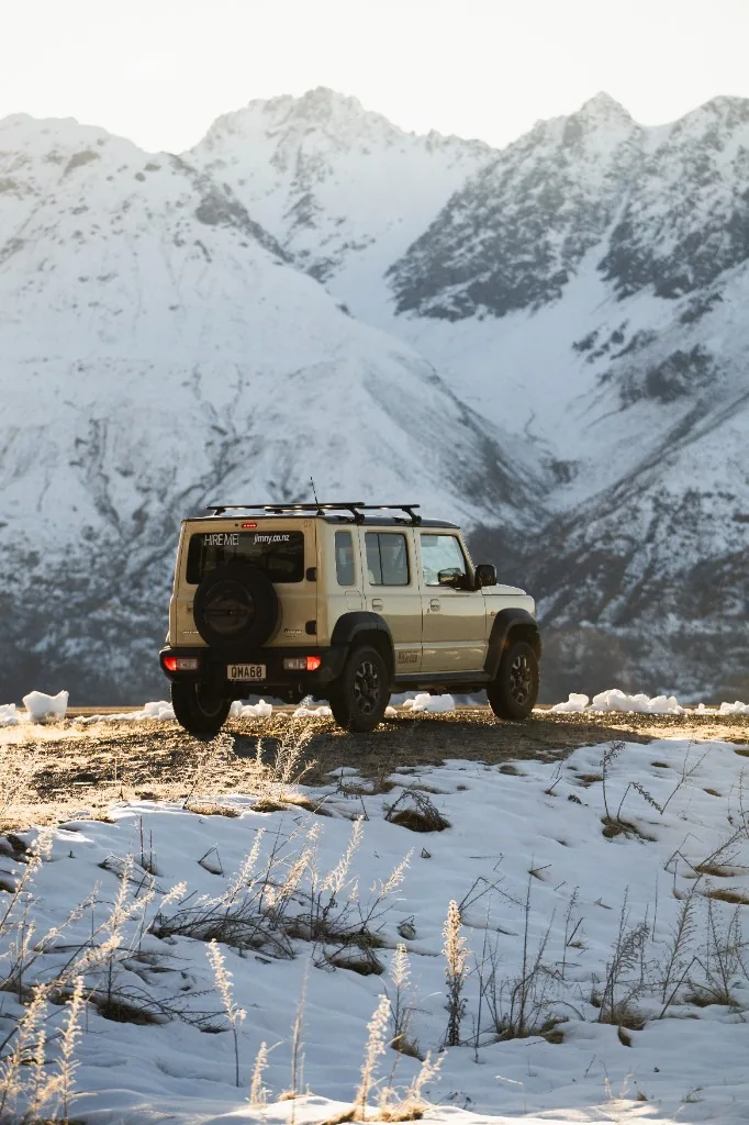 Tan Suzuki Jimny on a winding South Island road toward a snow-capped mountain.