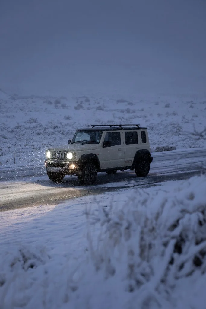 White Suzuki Jimny driving through heavy snow at night with headlights on, South Island winter.