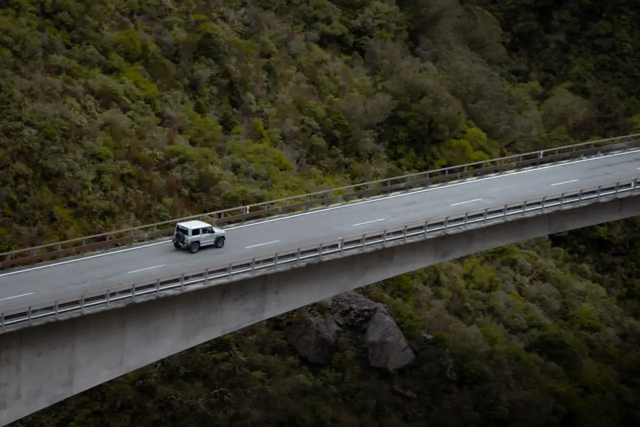 White Suzuki Jimny with rooftop tent in grassy clearing, mountains and trees. – Jimny Rentals.