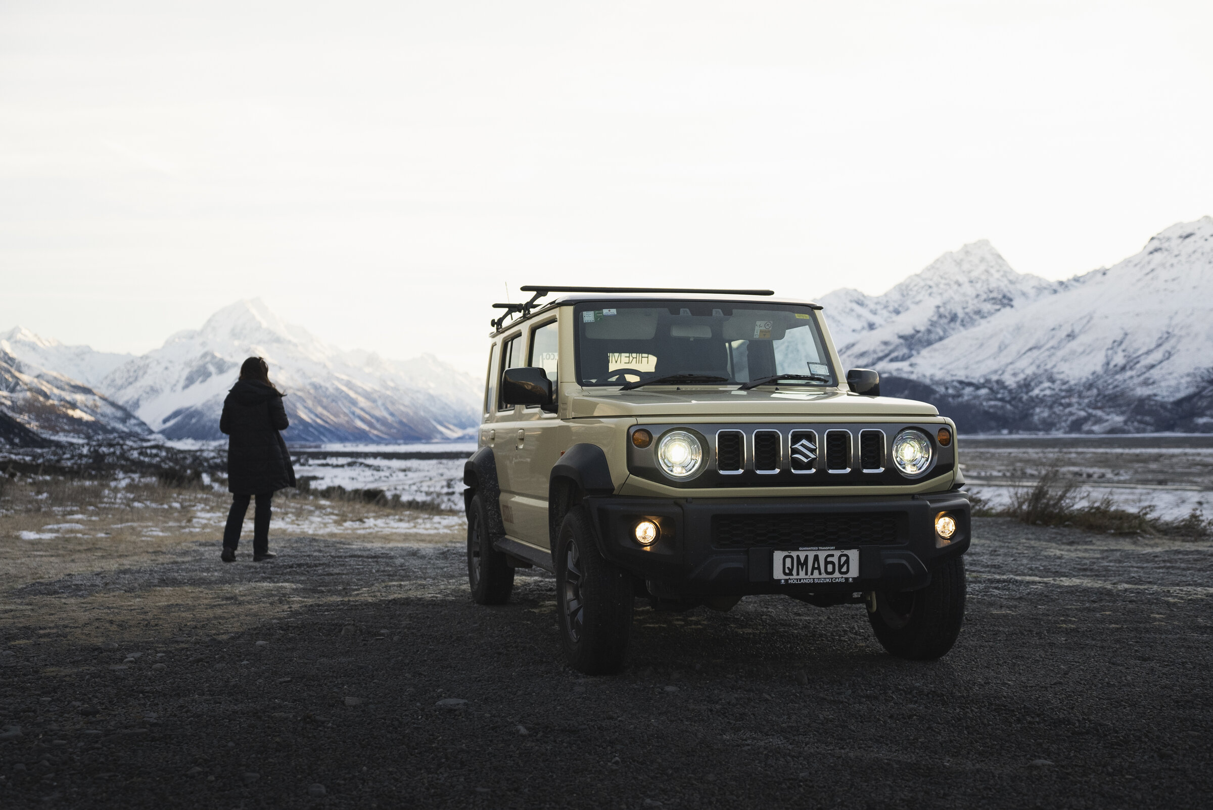 Cream Suzuki Jimny at a South Island alpine lookout, traveller looking toward snow-capped peaks