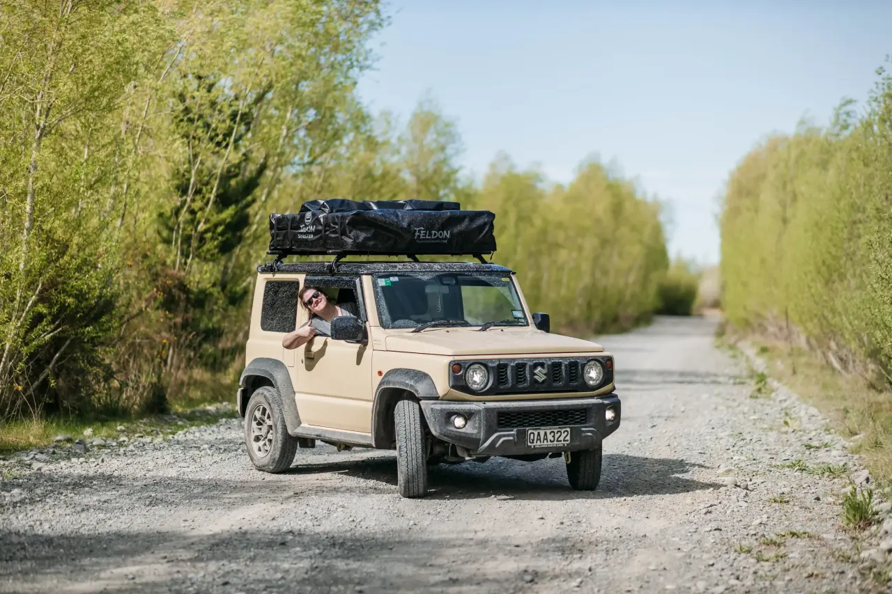 Cream Suzuki Jimny on a New Zealand gravel road, driver giving thumbs up out the window