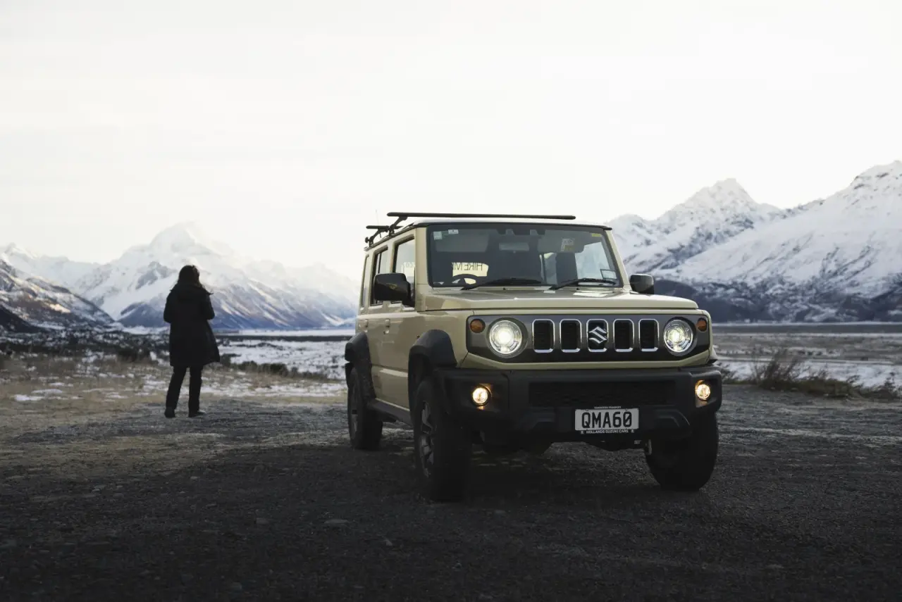 Tan Suzuki Jimny on a gravel road with snow-capped South Island mountains and a person enjoying the view.